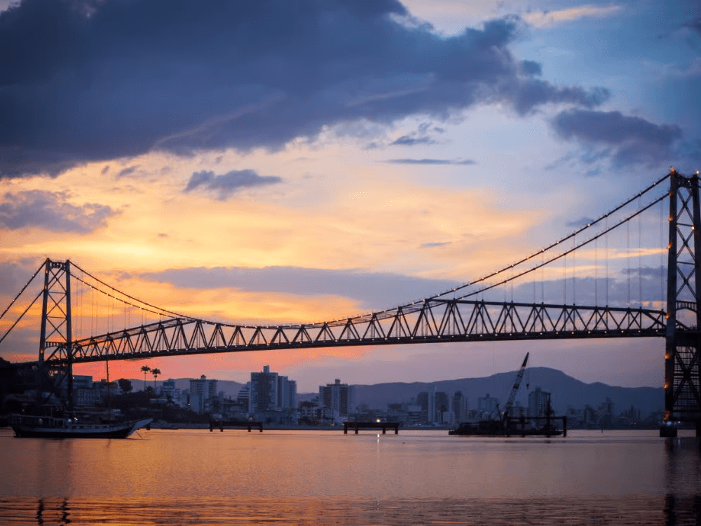 Vista da Ponte Hercílio Luz ao pôr do sol com a baía e parte da área urbana ao fundo, símbolo de Florianópolis. Tudo para mostra os melhores bairros para morar em Florianópolis.
