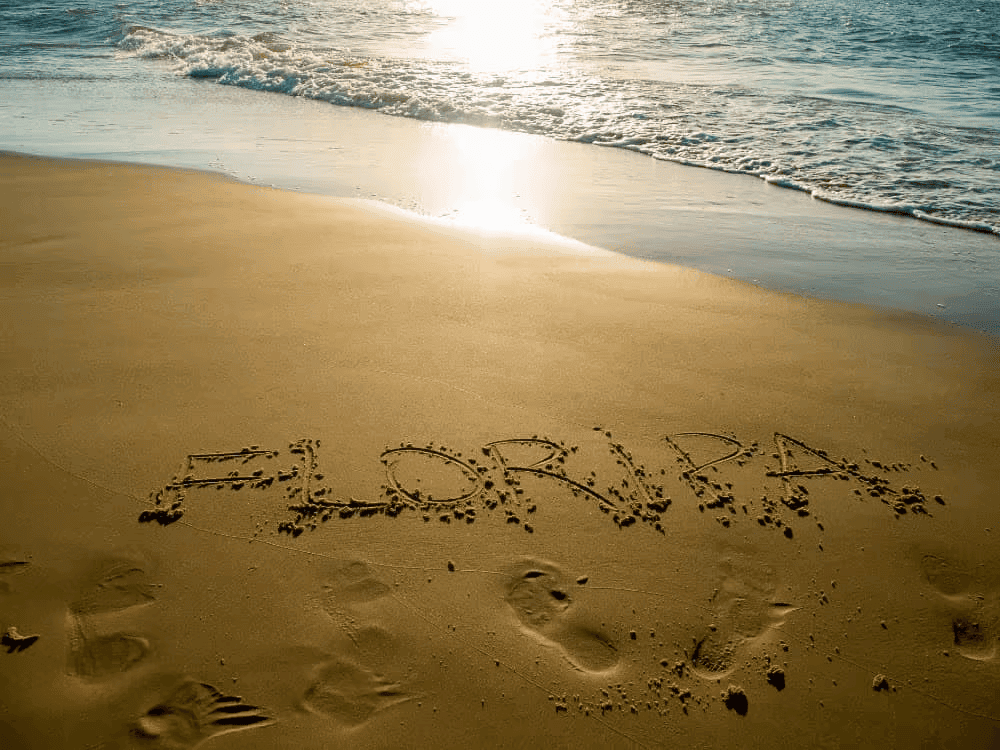 Faixa de areia com o nome Florianópolis escrito, ondas suaves e luz do sol refletindo no mar em um dia de praia.