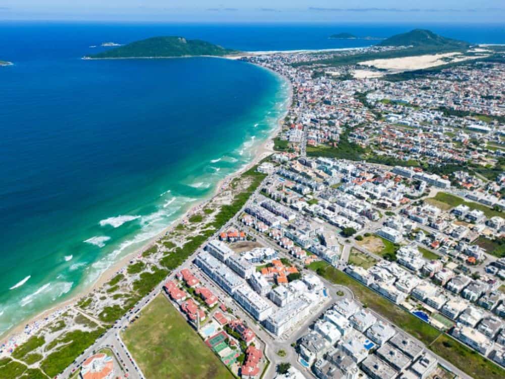 Vista aérea da praia de Jurerê, em Florianópolis, mostrando a faixa de areia, o mar azul com ondas suaves e a orla com casas e edifícios de alto padrão. Duas ilhas cobertas de mata aparecem no mar, e o céu está claro. Florianópolis é seguro e com qualidade de vida