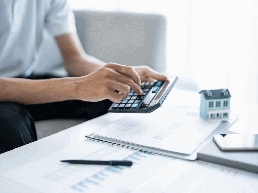 Pessoa utiliza calculadora ao lado de documentos e miniatura de casa para calcular parcelas do financiamento de imóvel usado.