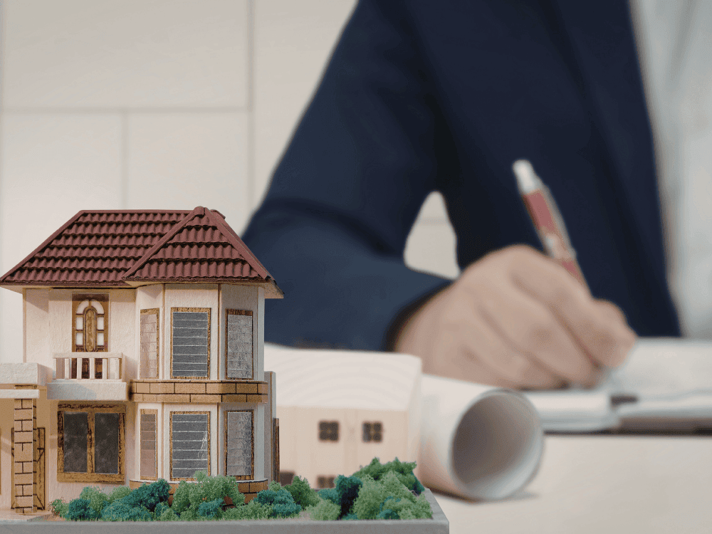 Maquete de uma residência sobre a mesa enquanto um profissional analisa documentos relacionados à compra de um imóvel usado.