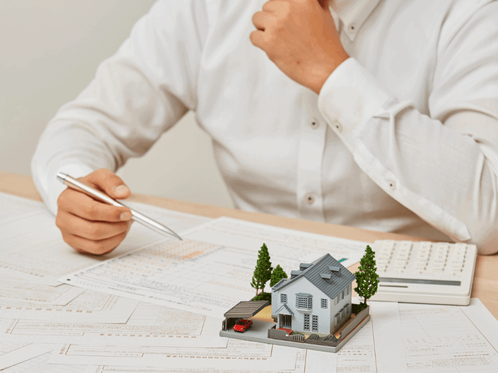 Profissional analisa documentos com maquete residencial e calculadora sobre a mesa durante avaliação do valor de um imóvel.