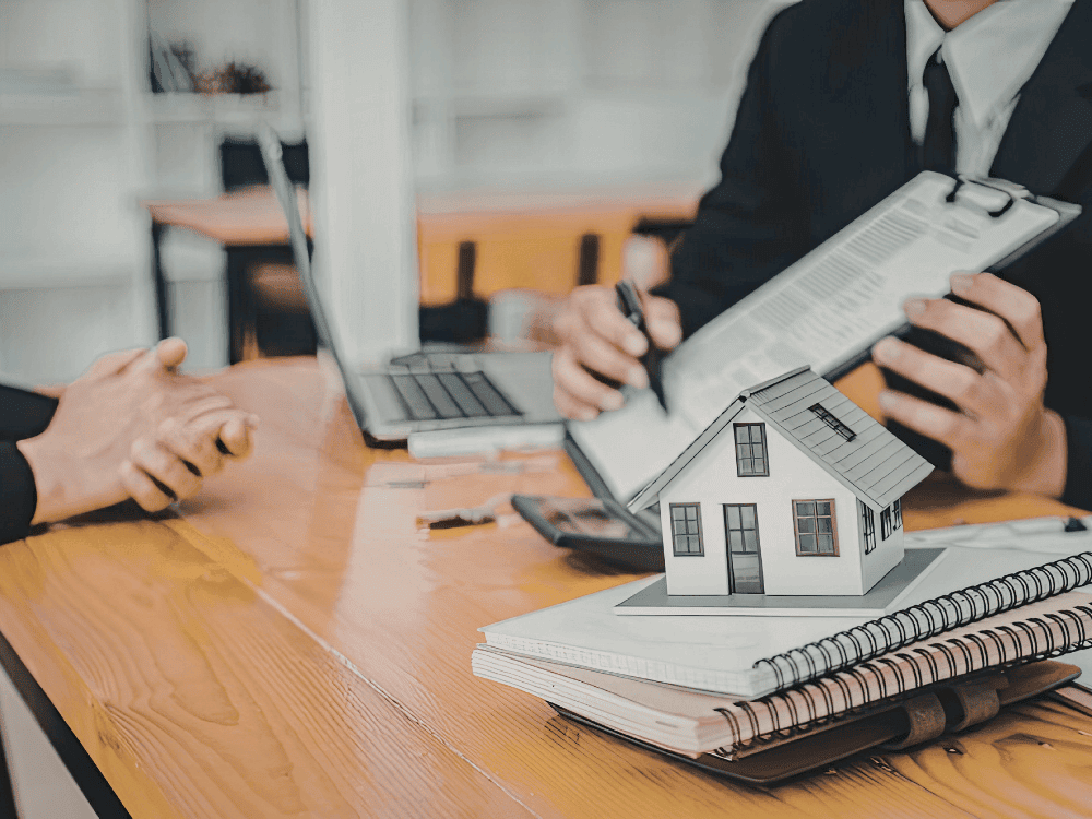 Consultor apresenta documentos ao cliente com maquete de casa sobre a mesa durante atendimento sobre valorização imobiliária.