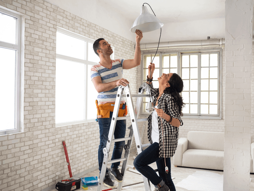 Casal realizando reparos em um imóvel, com ferramentas e instalação de luminária durante uma reforma para venda. Tudo para explicar se reformar para vender vale a pena.