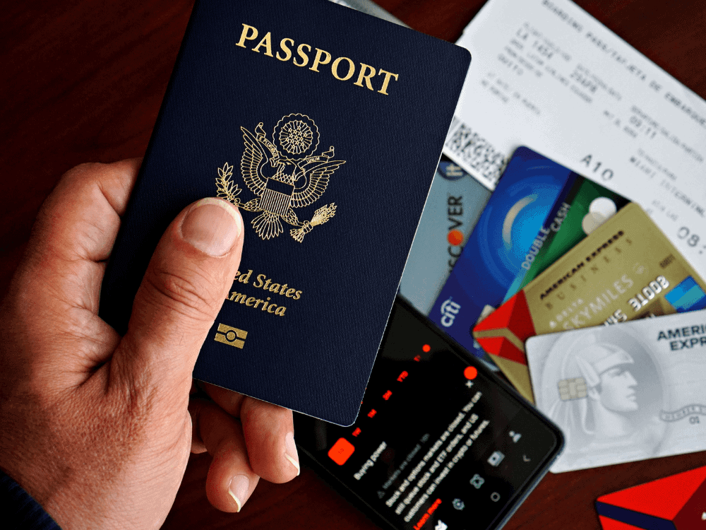 Mão segura um passaporte dos Estados Unidos sobre cartões bancários e um celular, representando a documentação de um comprador estrangeiro.